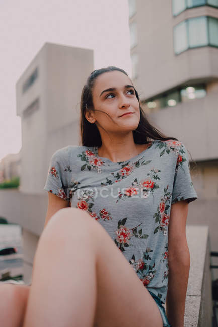 Ruhige, zufriedene junge Frau in kurzen Hosen und T-Shirt, die Sonne genießt, während sie auf einer Betonbrüstung sitzt und wegschaut — Stockfoto