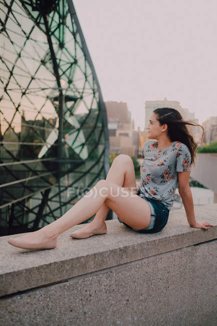 Ruhige, zufriedene junge Frau in kurzen Hosen und T-Shirt, die Sonne genießt, während sie auf einer Betonbrüstung sitzt und wegschaut — Stockfoto