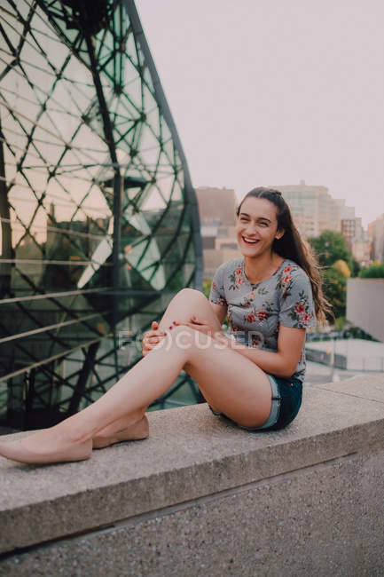 Спокойная девушка в шортах и футболке, наслаждаясь солнцем, сидя на бетонном парапете, глядя в камеру — Смотреть в камеру, город - Stock Photo