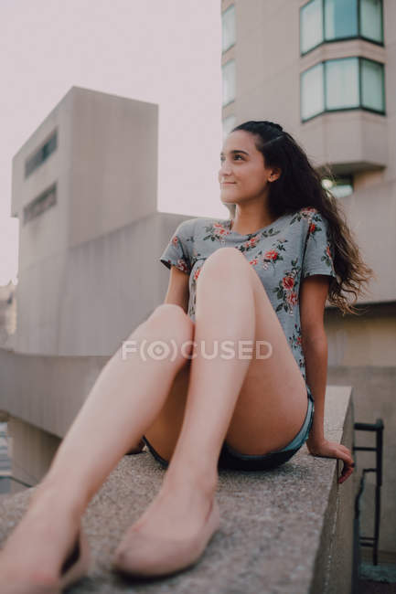 Спокойная девушка в шортах и футболке, наслаждаясь солнцем, сидя на бетонном парапете, отворачиваясь — стоковое фото