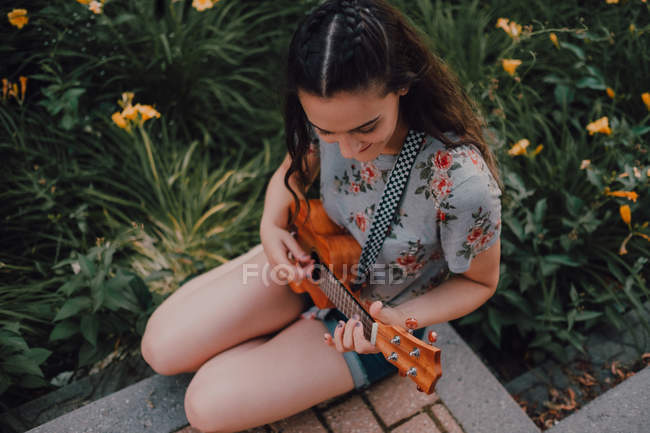 Smiling trendy casual young woman in t-shirt playing ukulele while sitting on pavement beside flowerbed — Stock Photo