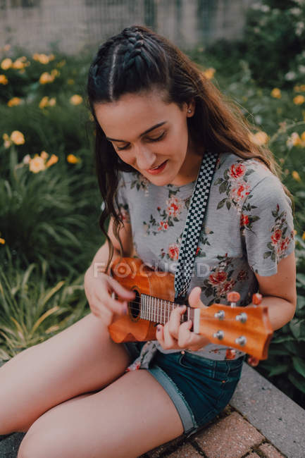 Усміхнена модна випадкова молода жінка в футболці грає укулеле, сидячи на тротуарі біля клумби — стокове фото