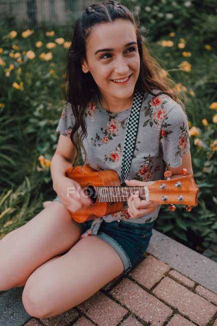 Smiling trendy casual young woman in t-shirt playing ukulele while sitting on pavement beside flowerbed — Stock Photo