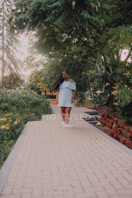 Noir jeune femme avec de longues tresses en robe hors épaule sur le chemin de béton dans le jardin en fleurs regardant caméra — Photo de stock