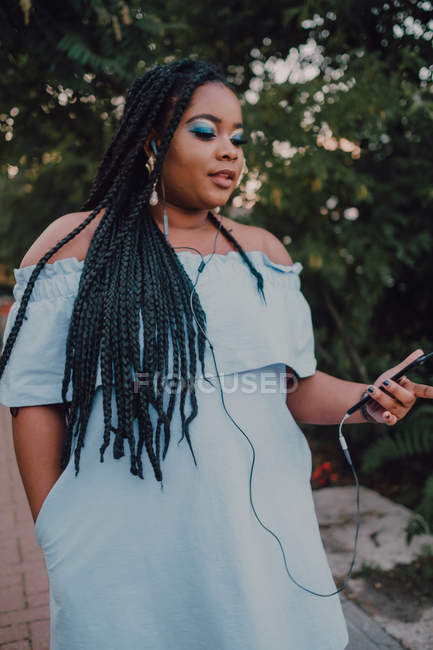 Jolie jeune femme noire avec un maquillage lumineux en robe off-shoulder écoutant de la musique sur smartphone avec écouteurs — Photo de stock