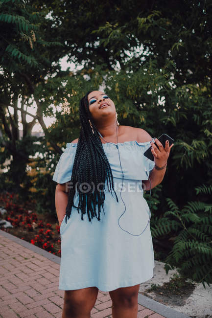 Atractiva joven negra con maquillaje brillante en vestido fuera del hombro escuchando música en el teléfono inteligente con auriculares - foto de stock
