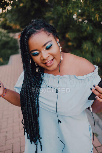 Attractive black young woman with bright make-up in off-shoulder dress listening to music on smartphone with earphones — Stock Photo