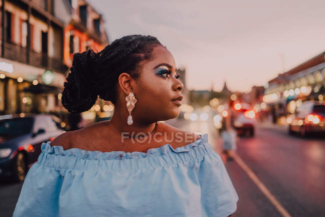 Attractive black young woman with bright makeup in off-shoulder dress standing on street on sunset, looking away — Stock Photo