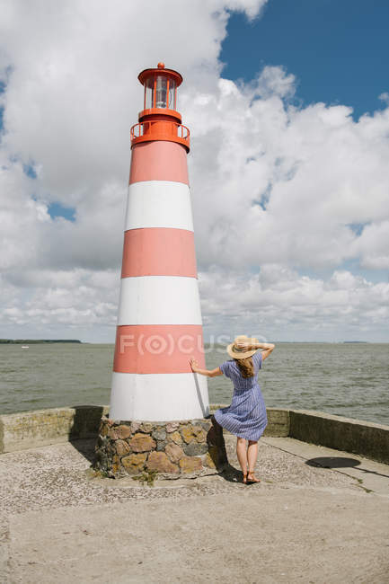 Vista trasera de la mujer en sombrero de paja y vestido ondulante de pie cerca del faro a rayas en la orilla en el día ventoso - foto de stock