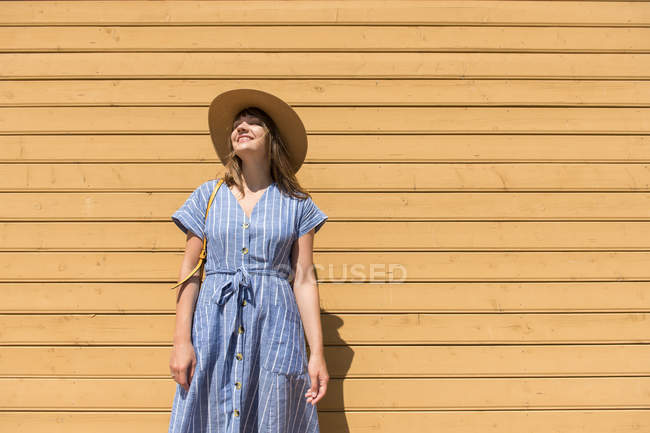 Smiling woman in straw hat and sundress standing with closed eyes in front of wooden wall — Stock Photo