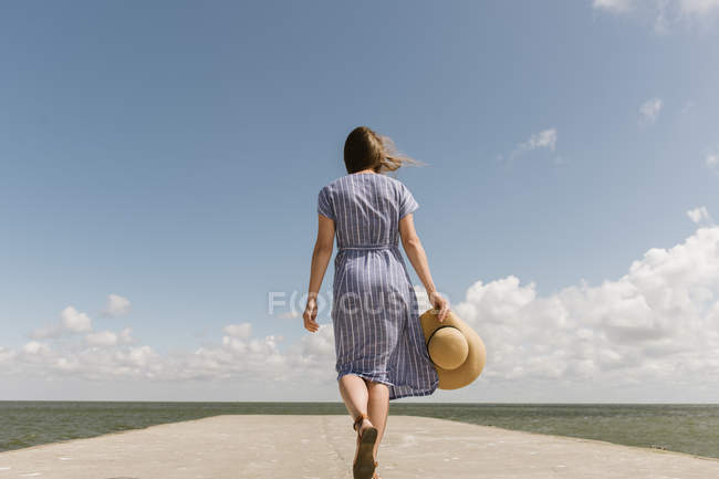 Visão traseira da mulher adulta em chapéu de palha e sundress no cais de concreto vazio no dia nublado — Fotografia de Stock