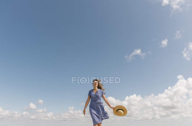 Взрослая женщина с развевающимися волосами и в сарафане ходит с соломенной шляпой в руке на облачном фоне неба — стоковое фото