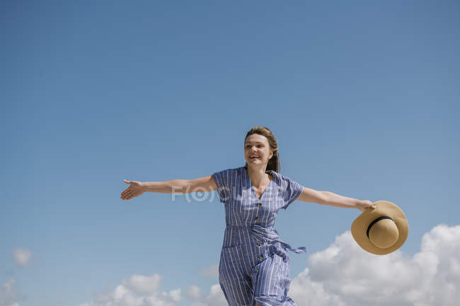Content adult woman with blowing hair and in sundress walking with straw hat in hand on cloudy sky background — Stock Photo