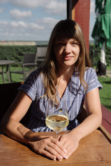 Відпочиваюча доросла жінка в одязі тримає келих на білому вині, сидячи за столом в альтанці в сонячний день — стокове фото