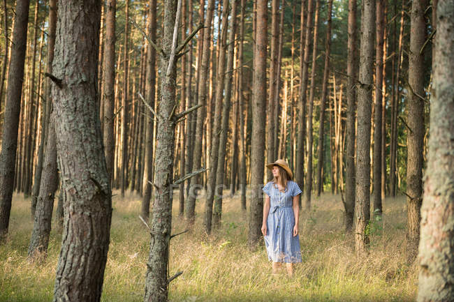 Щаслива доросла жінка в солом'яному капелюсі і сонячному одязі, що стоїть в лісі серед хвойних дерев в золотому сонячному промені — стокове фото