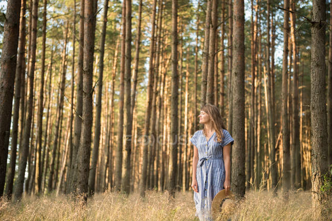 Heureuse femme adulte en chapeau de paille et robe de soleil debout dans la forêt parmi les conifères en rayon de soleil doré — Photo de stock