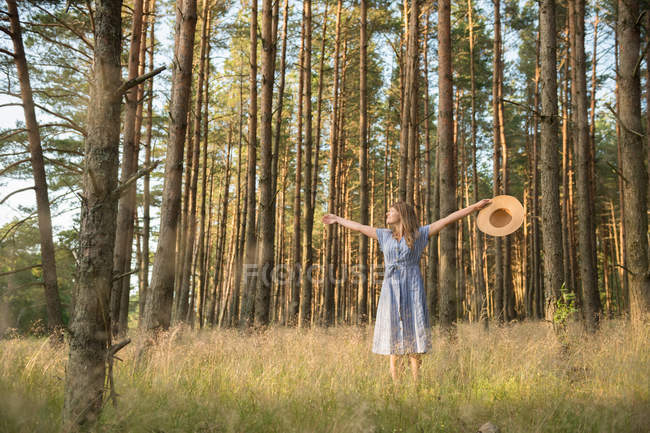 Happy adult woman in straw hat and sundress standing in forest among coniferous trees in golden sun ray — Stock Photo