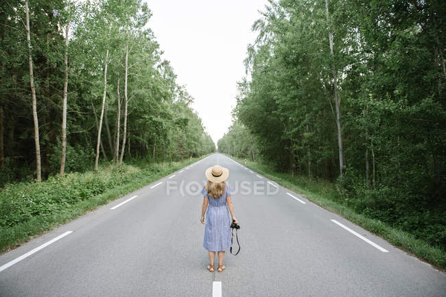 Visão traseira da mulher adulta em chapéu de palha e vestido com câmera em pé no meio do caminho de carro entre árvores verdes altas — Fotografia de Stock