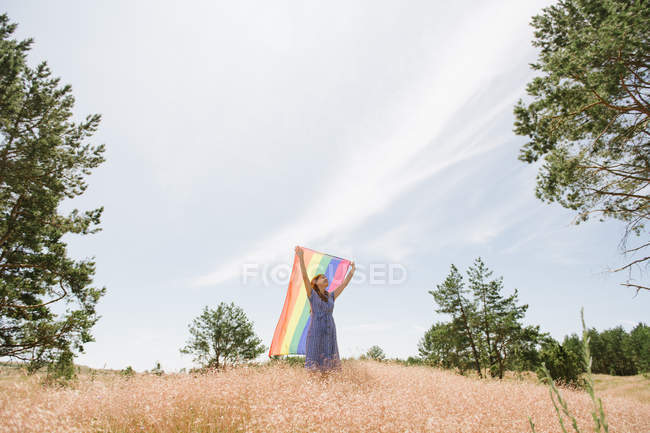 Вид на взрослую уверенную женщину в повседневной одежде с радужным флагом над головой в ветреный день — стоковое фото