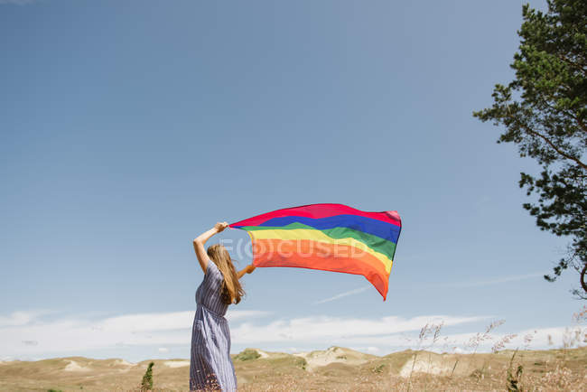 Side view of adult confident woman in casual dress carrying rainbow colored flag above head on windy day — Stock Photo