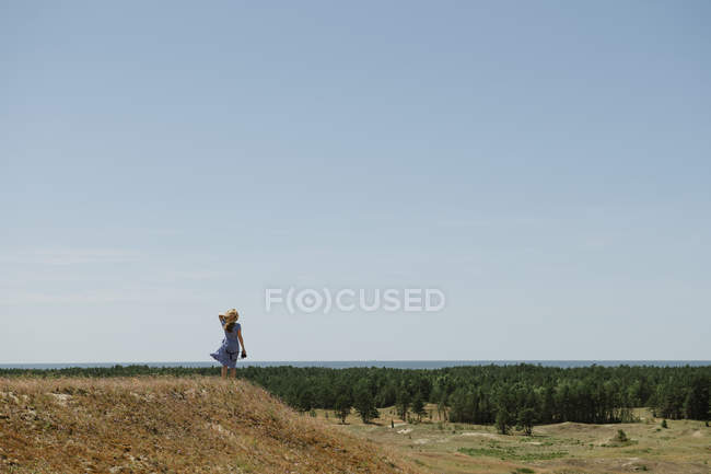 Vue arrière de la femme adulte en chapeau de paille et robe avec caméra debout sur la colline — Photo de stock