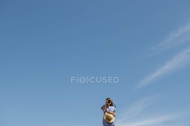 Mujer con sombrero de paja y vestido con cámara, de pie en un día soleado y ventoso con cielo azul claro en el fondo - foto de stock