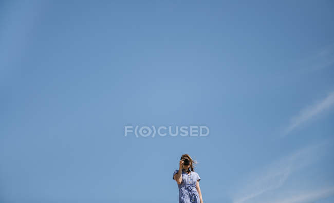 Donna in cappello di paglia e vestito con la macchina fotografica, in piedi in soleggiata giornata ventosa con cielo azzurro chiaro su sfondo — Foto stock