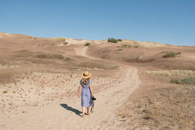 Rückansicht einer erwachsenen Frau in Strohhut und Kleid mit Kamera, die an sonnigen Tagen auf unbefestigter Straße zwischen trockenen staubigen Feldern spaziert — Stockfoto