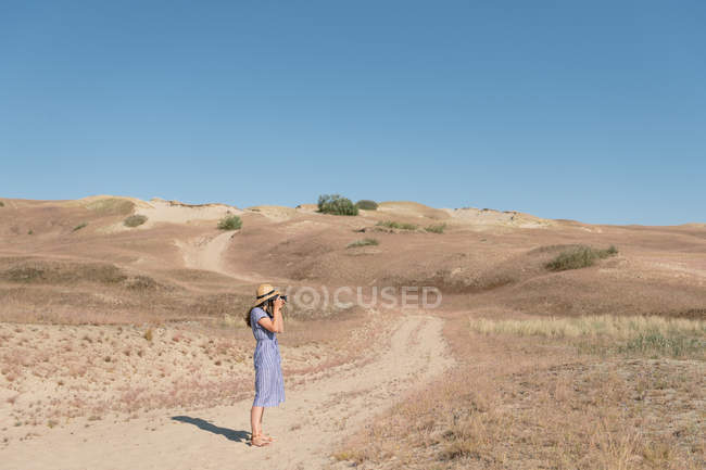 Femme adulte en chapeau de paille et robe avec caméra prenant des photos sur la route non pavée parmi les champs poussiéreux secs le jour ensoleillé — Photo de stock