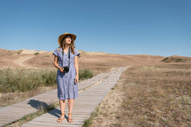 Доросла жінка в солом'яному капелюсі і в сукні з камерою, що стоїть на заміському шляху — стокове фото
