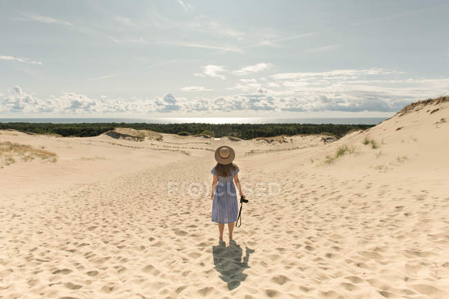 Visão traseira da mulher adulta em chapéu de palha e vestido com câmera andando na duna de areia da praia em um dia ensolarado — Fotografia de Stock