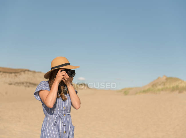Mulher adulta em chapéu de palha e vestido com câmera tirando foto na duna de areia da praia em um dia ensolarado — Fotografia de Stock