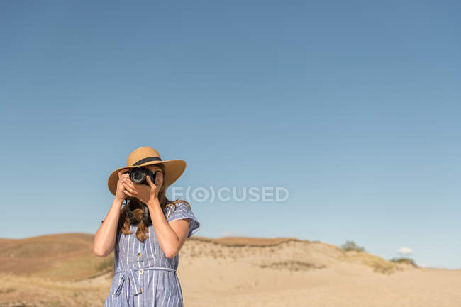 Adult woman in straw hat and dress with camera taking photo on sand dune of beach in a sunny day — Stock Photo