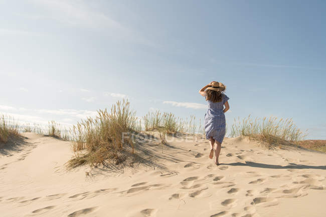 Mujer adulta en vestido casual y sombrero de paja caminando pasar un buen rato en la duna de arena en el día de verano caliente - foto de stock
