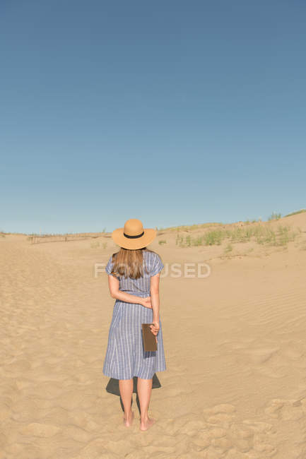 Mujer en vestido casual y sombrero de paja de pie con libro en la duna de arena en el caluroso día de verano - foto de stock