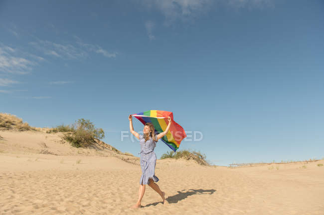 Adulte confiant femme en robe décontractée portant drapeau de couleur arc-en-ciel au-dessus de la tête sur les dunes de sable journée venteuse — Photo de stock