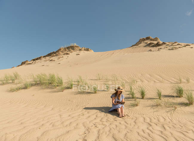 Nachdenkliche erwachsene Frau in lässigem Kleid und Strohhut liest Buch, während sie an heißen Sommertagen auf einer Sanddüne sitzt — Stockfoto