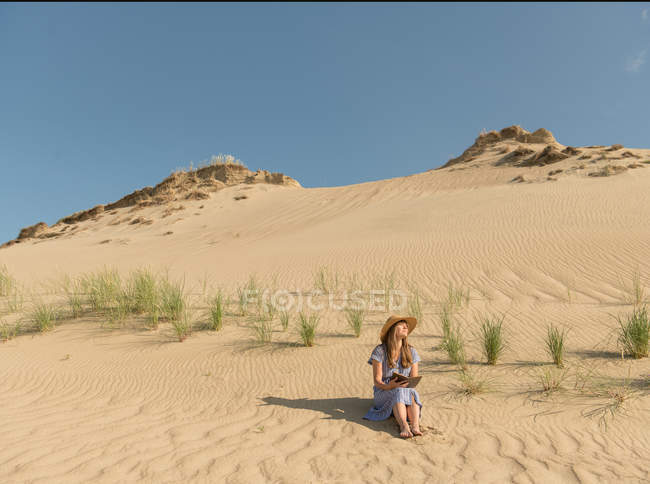 Ragionevole donna adulta in abito casual e cappello di paglia libro di lettura mentre seduto su dune di sabbia nella calda giornata estiva — Foto stock