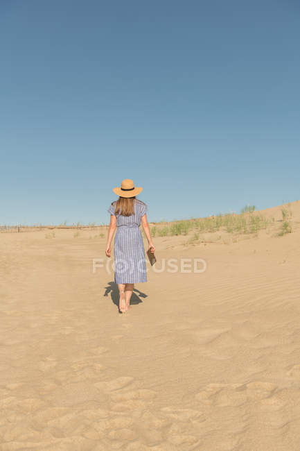 Mulher em vestido casual e chapéu de palha de pé com livro sobre duna de areia no dia quente de verão — Fotografia de Stock