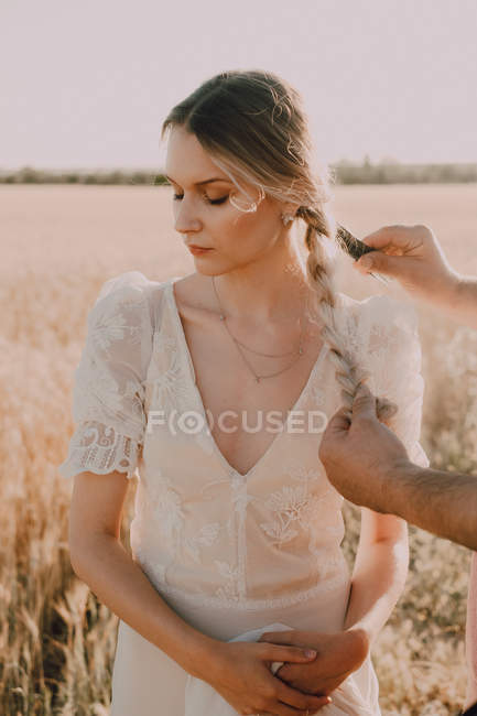 Shy attractive tender woman looking down folding hands while person combing braid hair in wheat field — Stock Photo