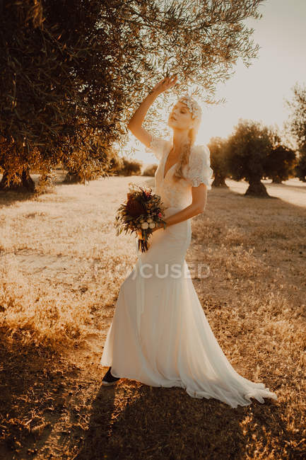 Vista lateral de la joven mujer tierna en vestido elegante luz blanca con gracia levantando la mano al árbol, sosteniendo flores a la luz del sol - foto de stock
