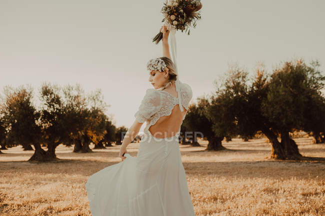 Вид ззаду на чуттєву ніжну жінку в мереживному платті, що тримає літню квітку в піднятих руках в полуденному сонці в саду дерев — стокове фото