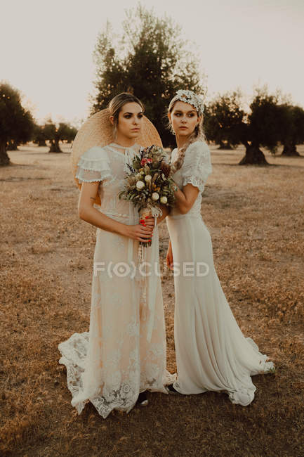 Mulheres pensativas bonitas em vestidos de casamento branco segurando buquês de flores e olhando ao longo — Fotografia de Stock