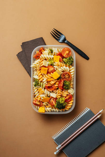 Vista dall'alto della scatola da pranzo in plastica con pasta, pomodori, formaggio e broccoli posizionati da forchetta in plastica nera e blocco note con matite su sfondo marrone — Foto stock