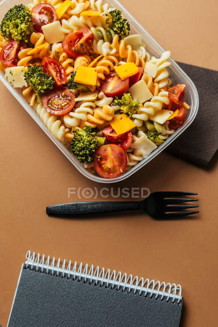 Lunchbox with pasta and plastic fork by notepad and pencils — Stock Photo