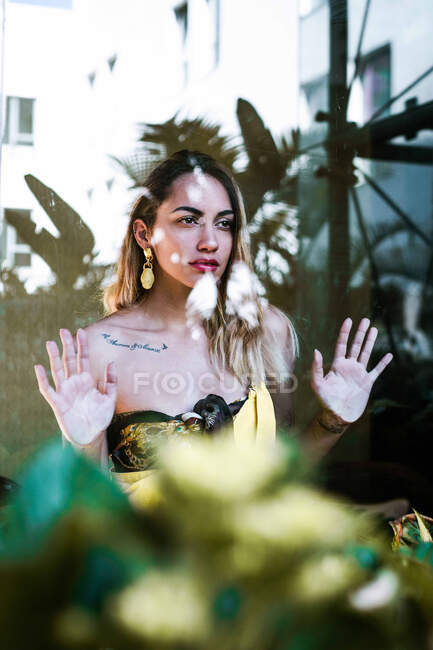 Jovem muito sério mulher confiante com maquiagem brilhante e tatuagem olhando para longe e tocando vidro com as mãos no fundo leve — Fotografia de Stock