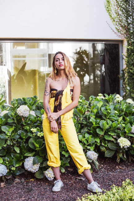 Junge hübsche Frau in modischer Kleidung steht im Garten mit Pflanzen und Blumen und blickt in die Kamera — Stockfoto
