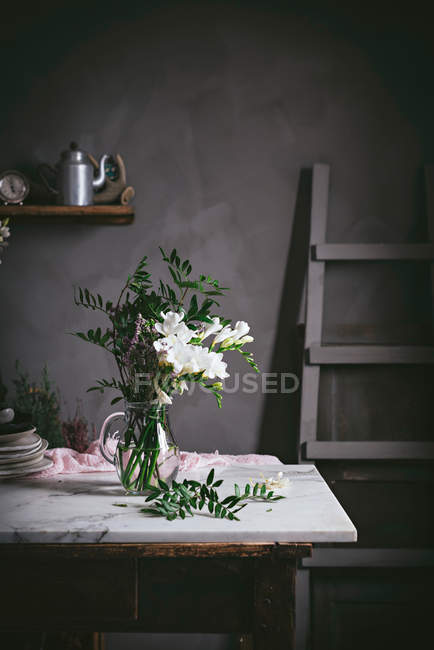 Florero de vidrio con ramo de flores blancas con hojas en la mesa de mármol junto a la pared oscura - foto de stock