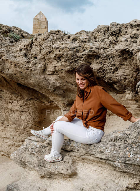 Vista lateral de la hermosa mujer feliz en vaqueros blancos sentados sobre piedras rocosas en el desierto. - foto de stock