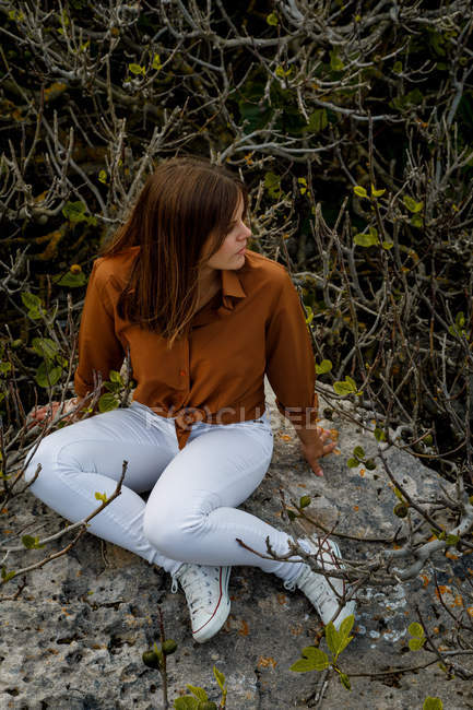 Cheerful beautiful woman sitting in big rock near bushes looking away — Stock Photo